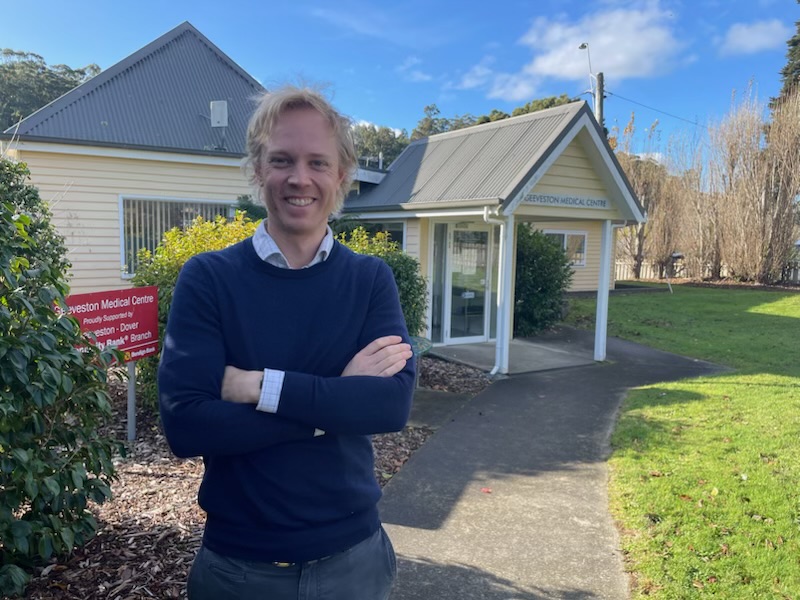 A person with light hair and a navy sweater stands smiling with arms crossed in front of a small building marked “Geeveston Medical Centre” on a sunny day. Green shrubs and a red information sign are visible.