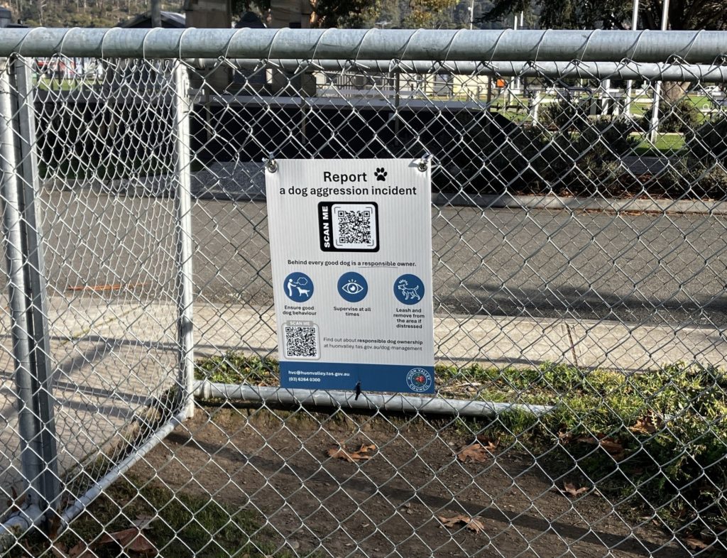 A sign attached to a chain-link fence reads, Report a dog aggression incident with QR codes and icons, located near a sidewalk and grassy area. Sunlight casts shadows on the ground and fence.