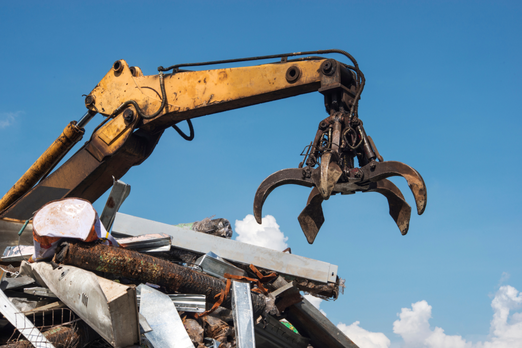 A yellow industrial claw crane lifts scrap metal and debris at a junkyard under a clear blue sky with scattered clouds.