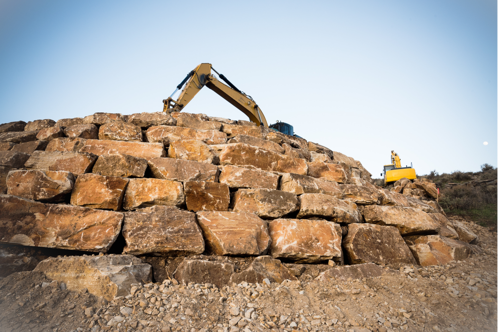 A large excavator works atop a mound of stacked, rough-cut stone blocks, with a clear sky and some vegetation visible in the background.