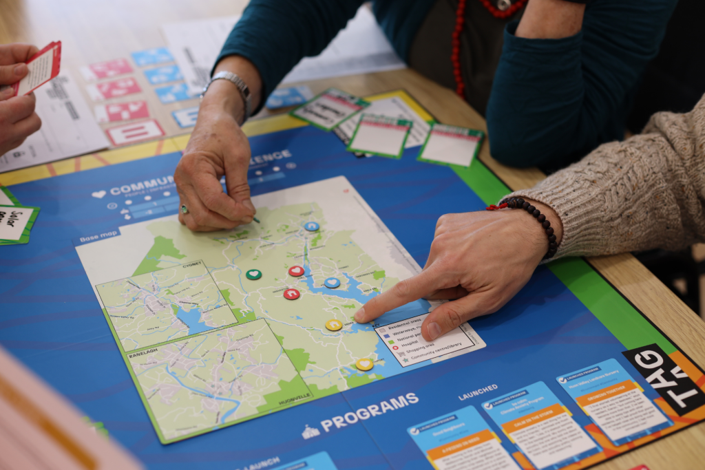 Two people play a strategy board game involving a map, colored tokens, and cards. One person points to a spot on the map, while the other prepares to move a piece. Various cards are visible around the board.