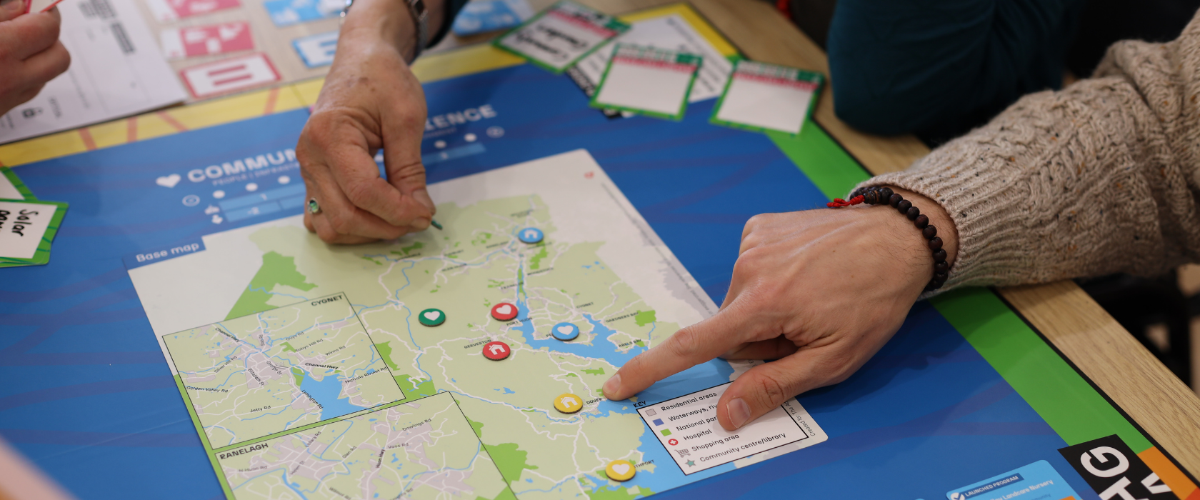 Two people play a strategy board game involving a map, colored tokens, and cards. One person points to a spot on the map, while the other prepares to move a piece. Various cards are visible around the board.
