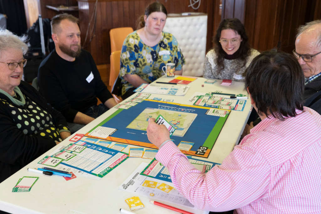 Six people are sitting around a table playing a board game. The table is covered with cards, game pieces, and a colorful board. The group appears to be engaged and enjoying the activity together.