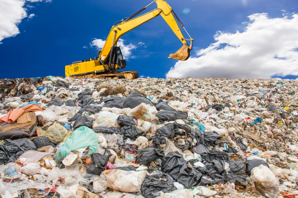 A yellow excavator sits atop a large heap of mixed waste and plastic bags at a landfill under a bright blue sky with scattered clouds.