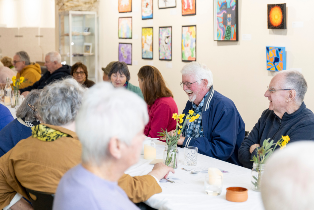 A group of older adults sit around a table with flowers, laughing and talking in a brightly lit art gallery decorated with colorful paintings.