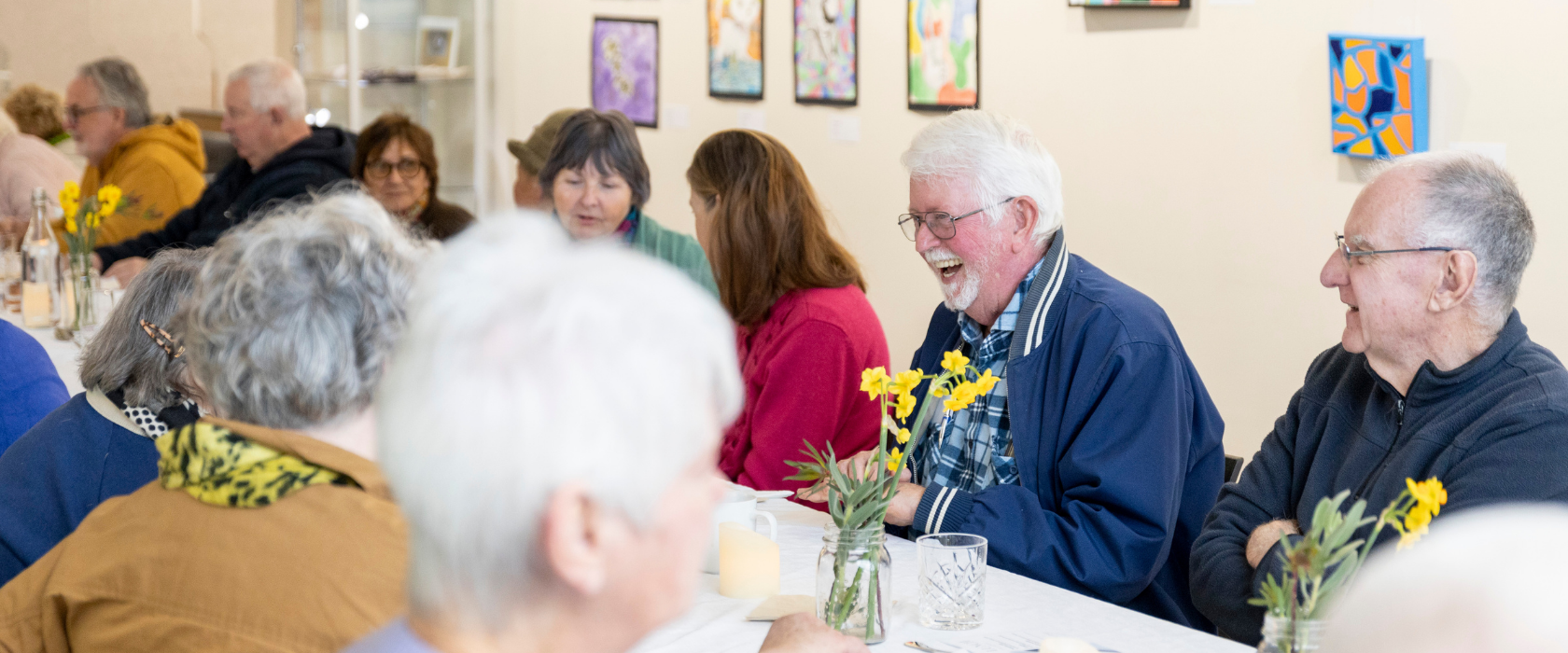 A group of older adults sit around a table with flowers, laughing and talking in a brightly lit art gallery decorated with colorful paintings.