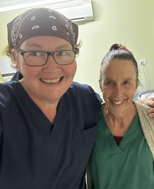 Two women in medical scrubs smile at the camera. One wears glasses and a bandana, while the other has her hair pulled back. They appear to be in a clinical or hospital setting with light-colored walls.