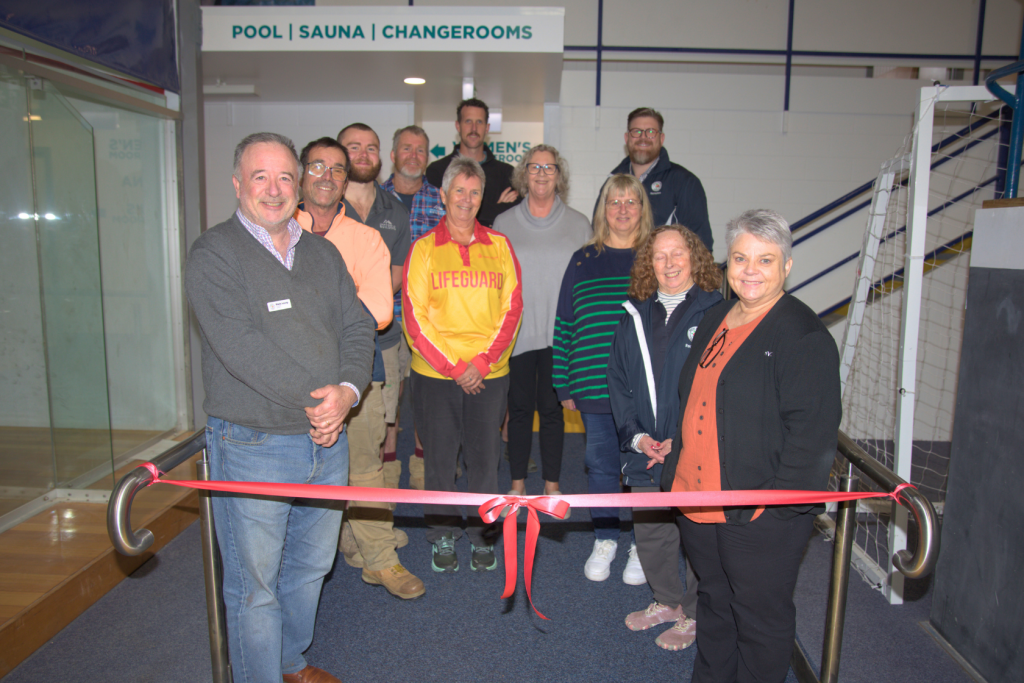 A group of people stand behind a red ribbon at an indoor facility, smiling at the camera. Signs in the background indicate directions to the pool, sauna, and changerooms. Some are dressed casually; one wears a lifeguard shirt.