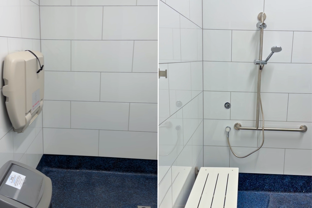 A public restroom with white tiled walls, featuring a baby changing station, a trash bin, a mounted showerhead, a grab bar, and a foldable bench.