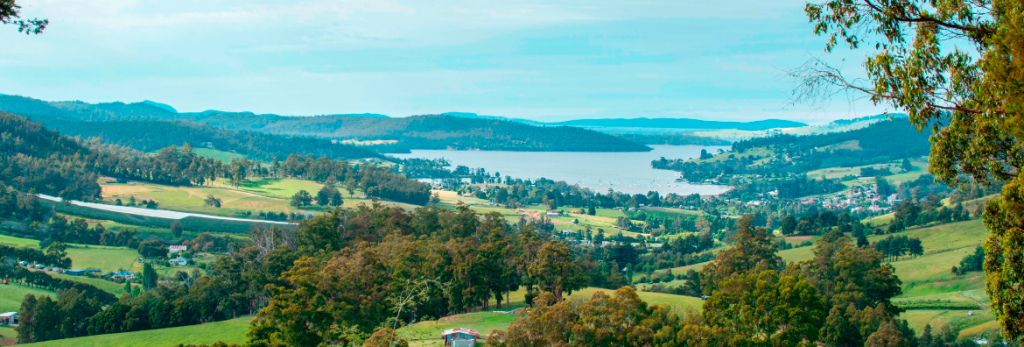 A panoramic view of a lush, green valley with scattered houses, dense trees, and rolling hills, leading to a calm blue river or lake under a clear sky.