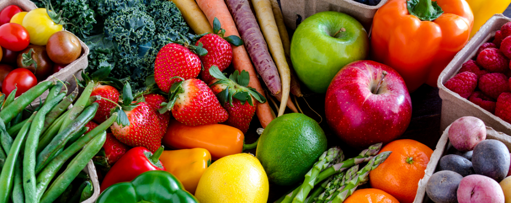 A colorful assortment of fresh fruits and vegetables, including apples, strawberries, citrus, green beans, asparagus, bell peppers, kale, carrots, raspberries, tomatoes, and plums, arranged closely together.