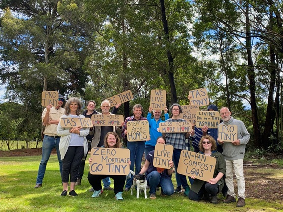A group of people outdoors hold cardboard signs supporting tiny houses and less house, more home messages. Signs include “PRO TINY,” “LIVE TINY,” and “UN-BAN COMPOST LOOS.” Trees and grass are in the background.