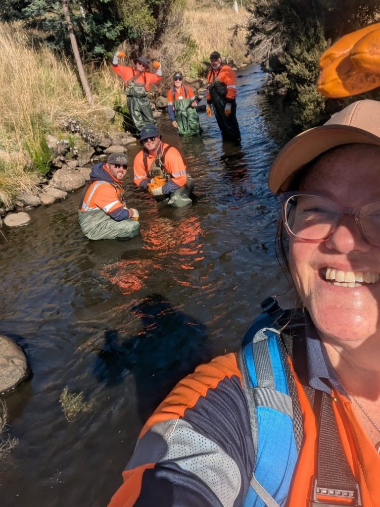 A group of people wearing orange shirts and waders stand in a shallow creek surrounded by grass and trees. One person in the foreground takes a smiling selfie. The group appears to be doing fieldwork or research.
