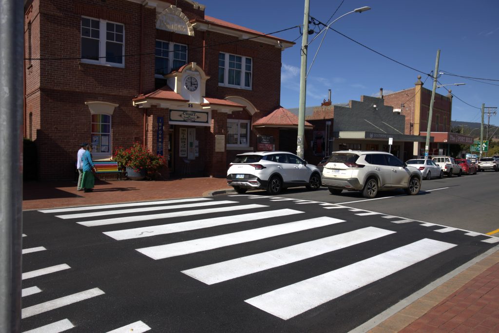 A zebra crossing leads to a brick building with a sign above the entrance. Several cars are parked along the street, and two people are walking on the sidewalk. The sky is clear and blue.