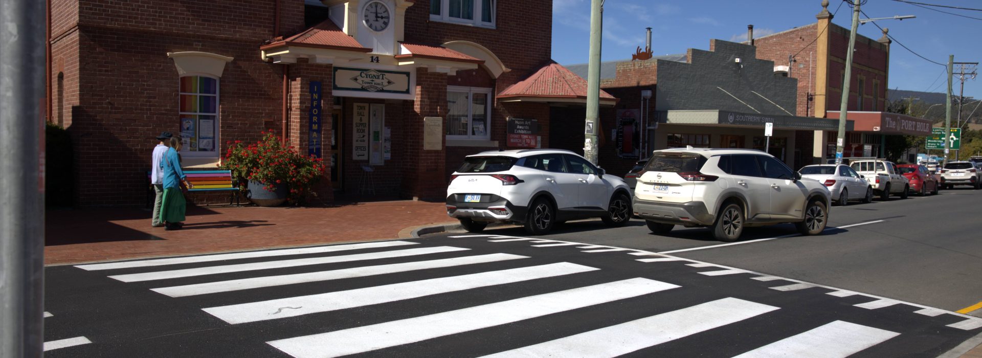 A zebra crossing leads to a brick building with a sign above the entrance. Several cars are parked along the street, and two people are walking on the sidewalk. The sky is clear and blue.