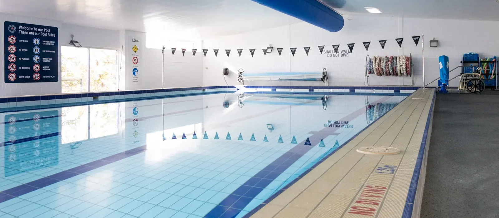Indoor swimming pool with clear blue water, lane markers, and safety signs on the wall. Poolside tiles read NO DIVING and there are swimming aids and equipment neatly stacked along the side.