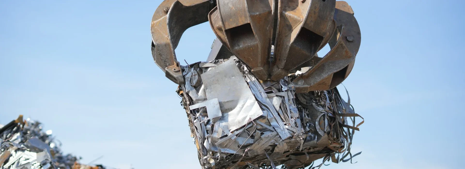 A large industrial claw lifts a bundle of scrap metal in a recycling yard, with piles of metal debris visible in the background against a clear blue sky.