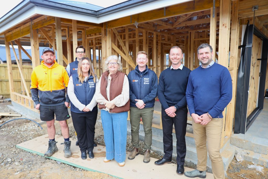 A group of seven people stand smiling in front of a partially constructed wooden house frame at a building site. Some wear work uniforms, others are dressed in business attire.