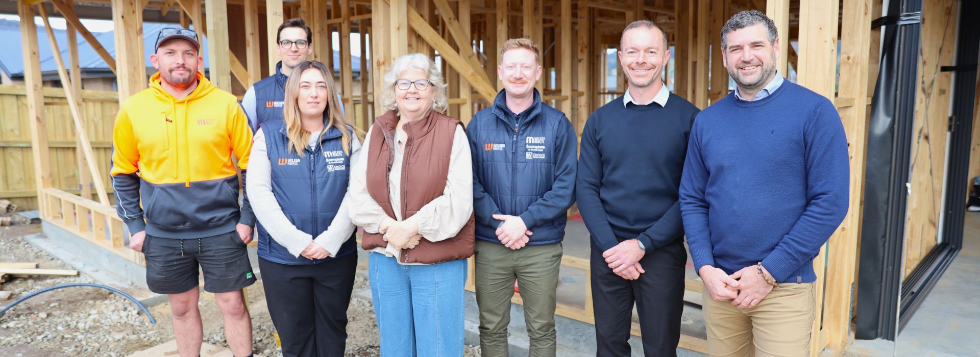 A group of seven people stand smiling in front of a partially constructed wooden house frame at a building site. Some wear work uniforms, others are dressed in business attire.