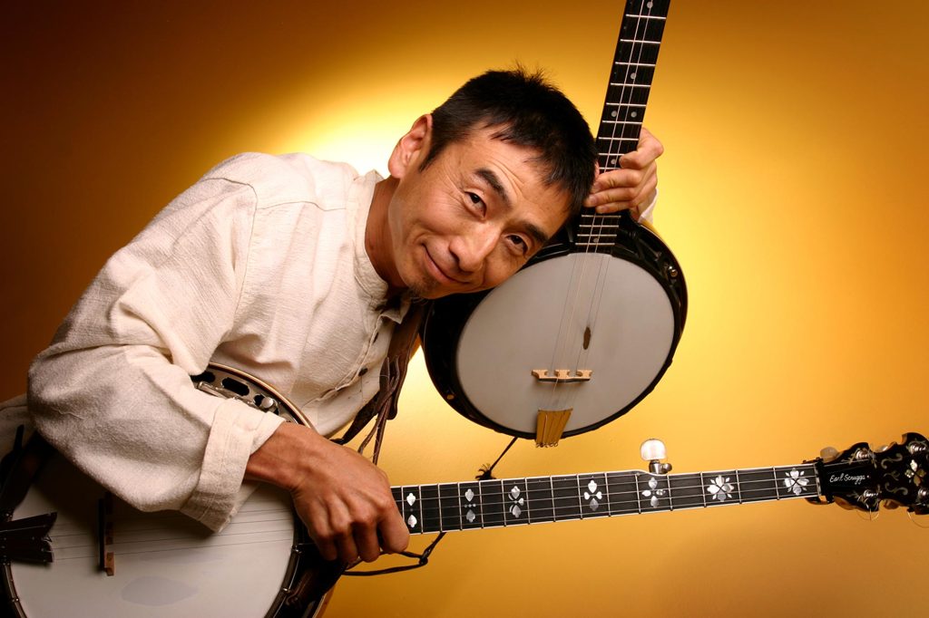 A man in a cream shirt smiles while holding and playing two banjos against a warm yellow background.