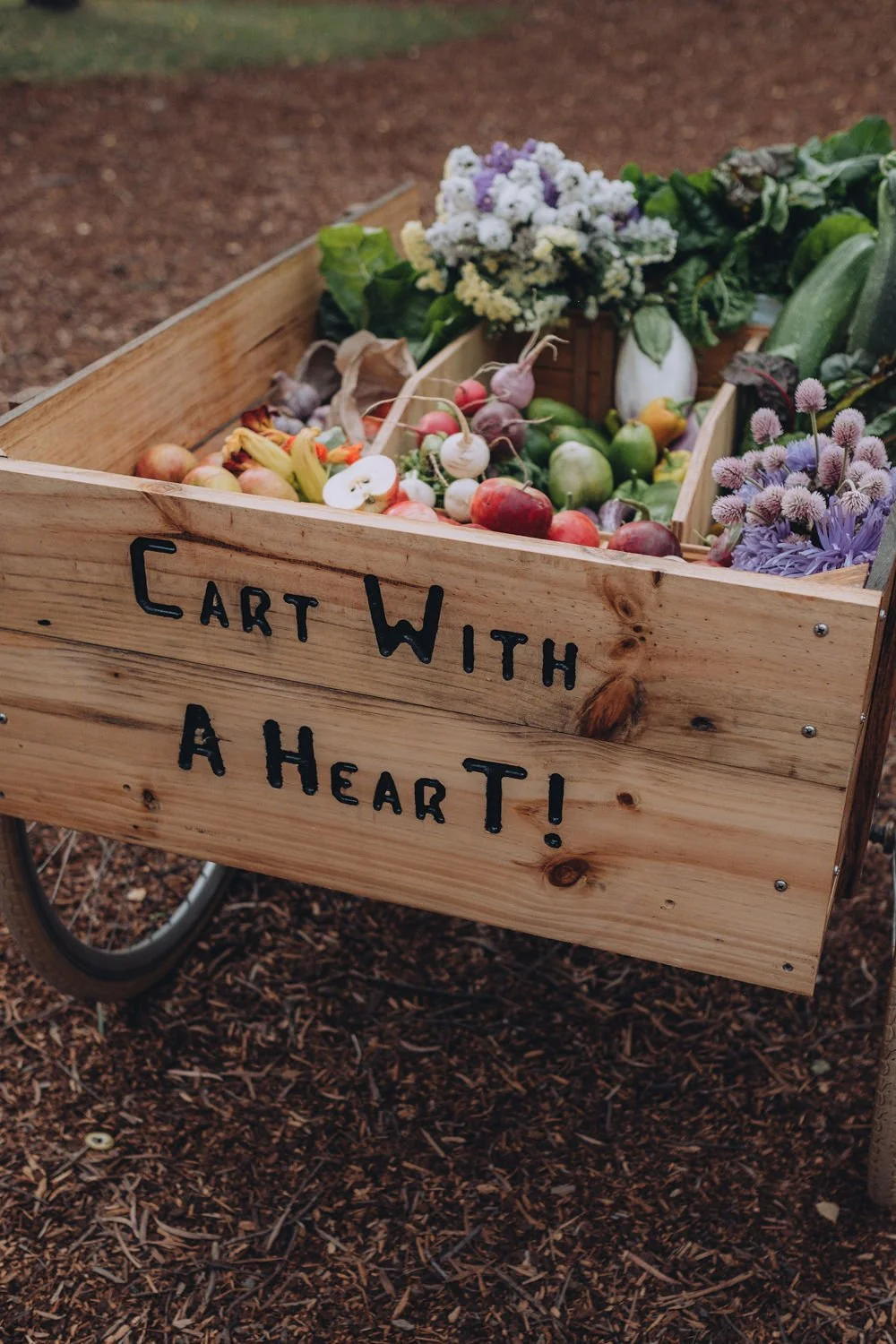 A wooden cart with Cart With A Heart! written on it is filled with assorted fresh vegetables, fruits, and flowers, sitting on mulch outdoors.