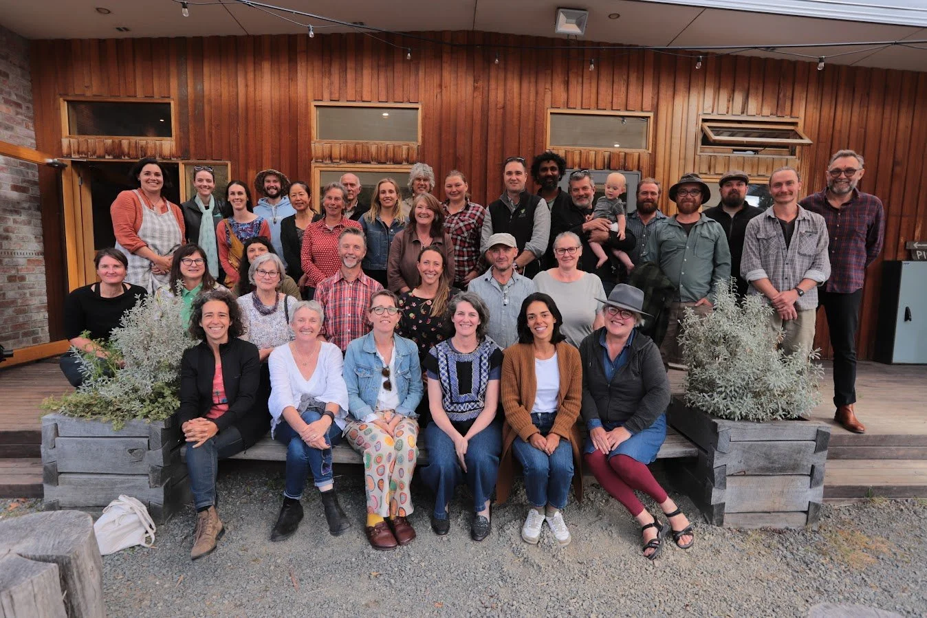 A group of about 30 adults poses together outside a wooden building, smiling at the camera. Some are seated in front, while others stand behind them. There are plants in front of the group and gravel on the ground.