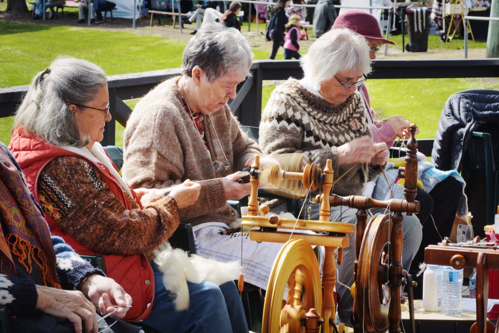 Three elderly women sit outdoors, spinning wool using spinning wheels. They wear sweaters and glasses, concentrating on their work. Other people and market stalls are visible in the background on a grassy area.