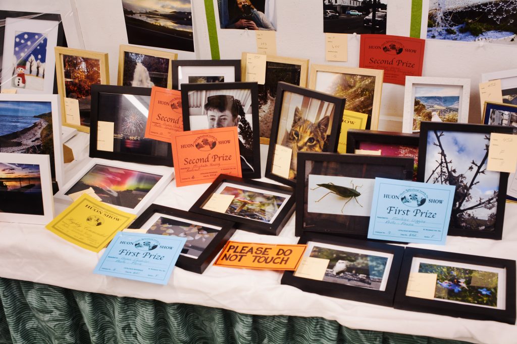A display table with framed photographs, colorful award certificates for first and second prizes, and a βPLEASE DO NOT TOUCHβ sign. The photos include landscapes, animals, and people.