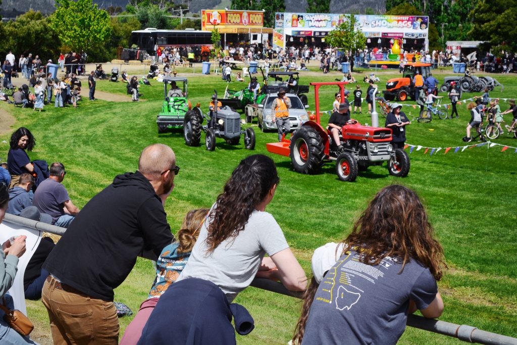 People watch a parade of vintage tractors driving across a grassy field at an outdoor event, with food stalls, crowds, and trees in the background under a sunny sky.