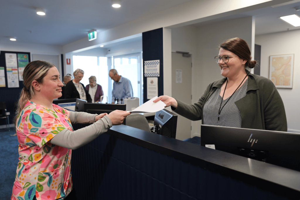 A woman in colorful scrubs hands paperwork to a receptionist across a counter, with several people waiting in line in the background of a modern office setting.