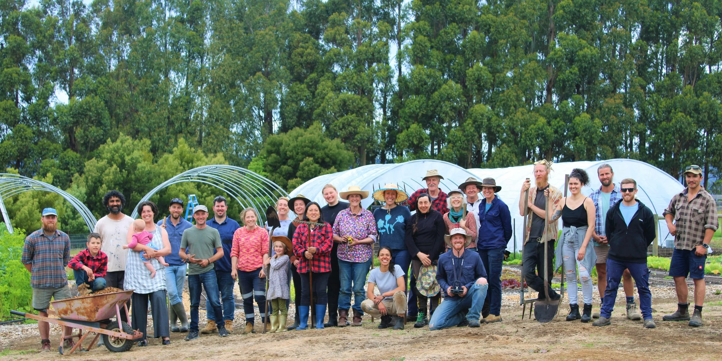 A large group of people pose outdoors on a farm, some holding tools. Greenhouses and tall trees are in the background. The group includes adults, children, and a dog.
