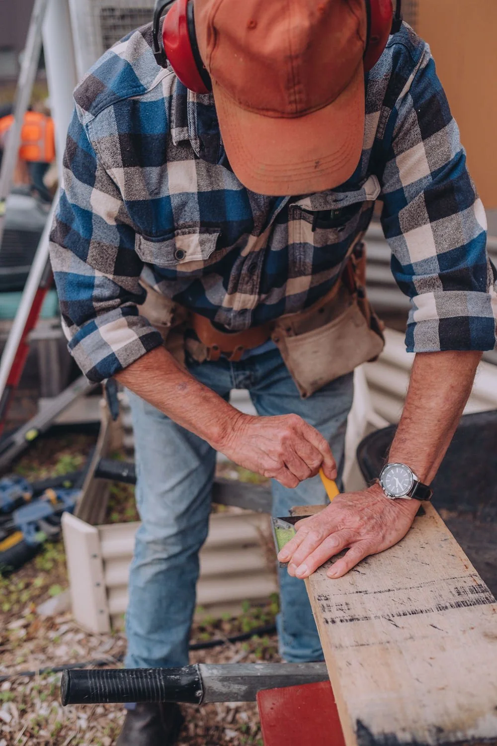 A person wearing a plaid shirt, jeans, and a cap uses a measuring tool on a wooden plank outdoors. Tools and equipment are visible in the background.