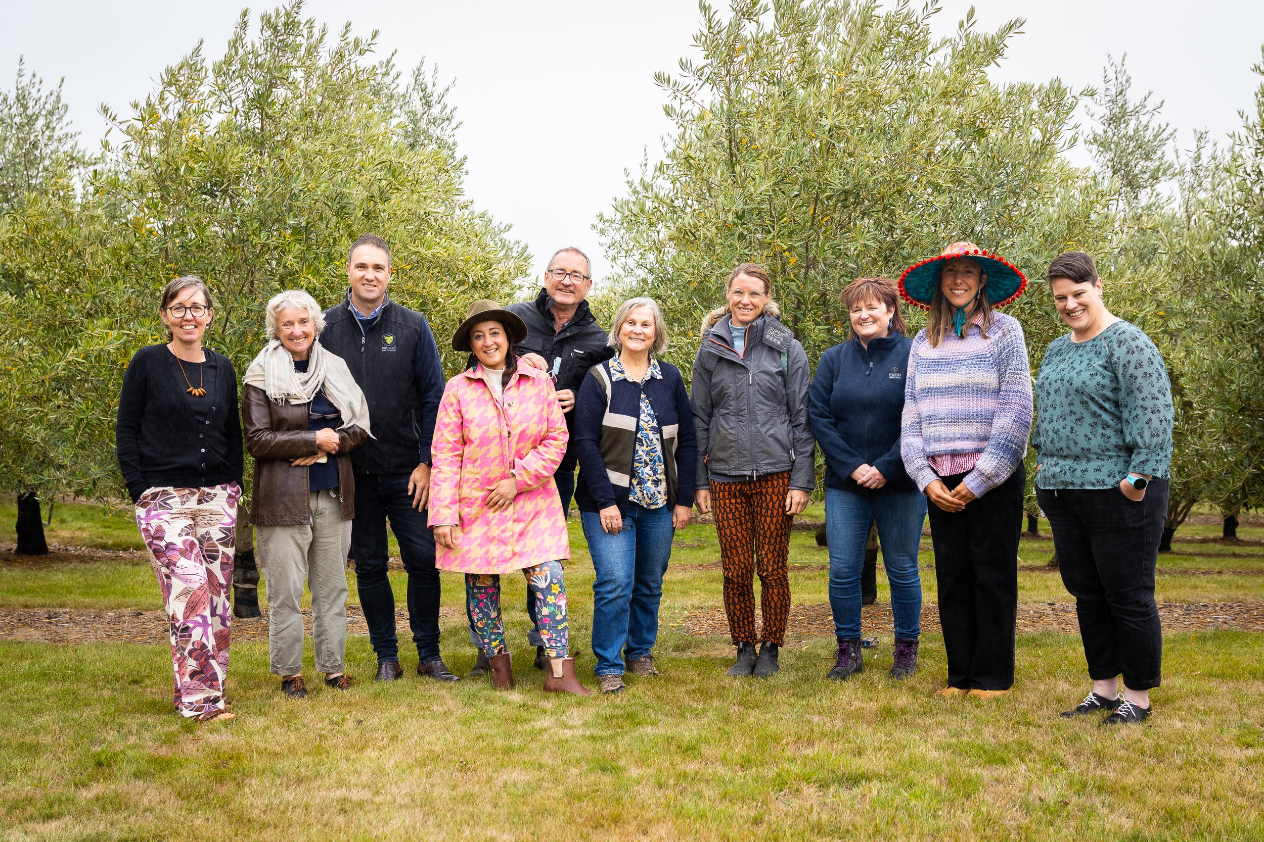 A group of ten adults stands smiling in a grassy field among trees. Several wear colorful clothing and hats. The weather appears overcast.
