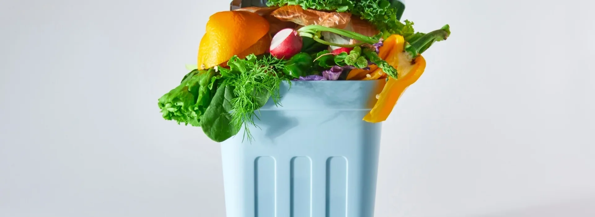 A light blue trash bin filled with assorted food scraps, including vegetables, greens, orange peels, and bread, against a plain white background.