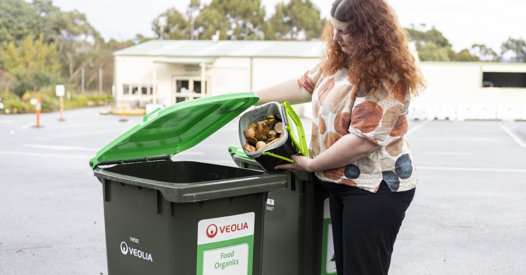 A woman stands outside, lifting the lid of a green food organics bin and emptying a container of food scraps into it. The bin is labeled with the Veolia logo and Food Organics.
