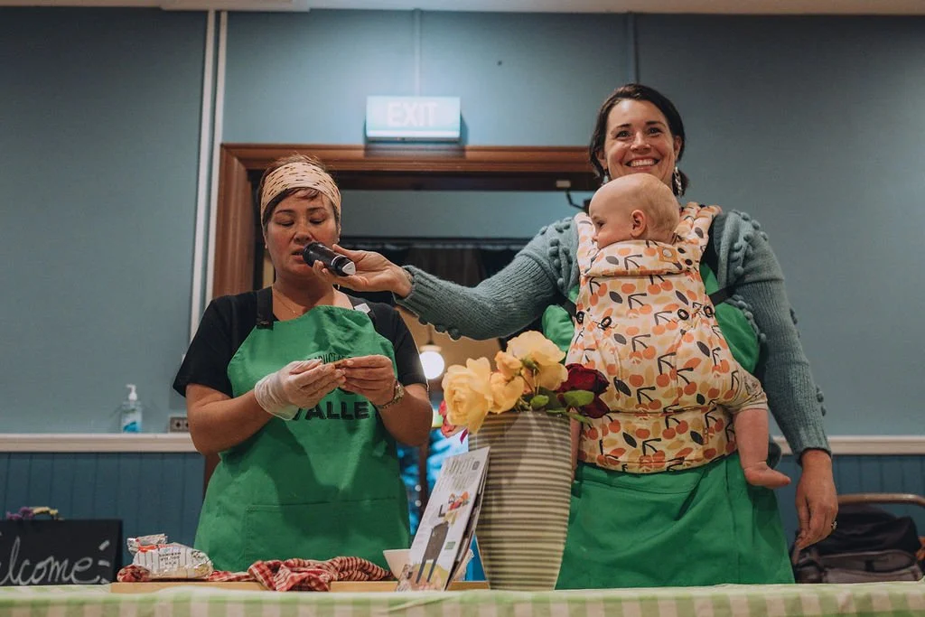 Two women stand at a table with flowers. One holds a microphone and wears a baby in a carrier. The other, wearing gloves, speaks into the mic. Both wear green aprons.