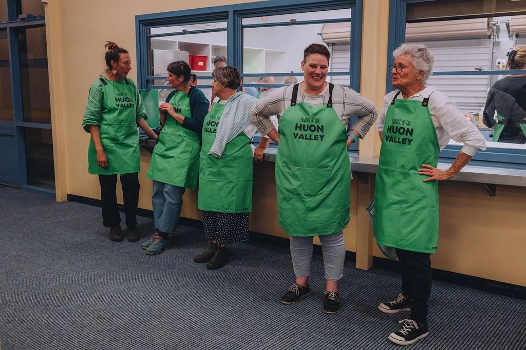Five people wearing green Huon Valley aprons stand and chat in a room with blue trim and a counter, appearing relaxed and friendly.