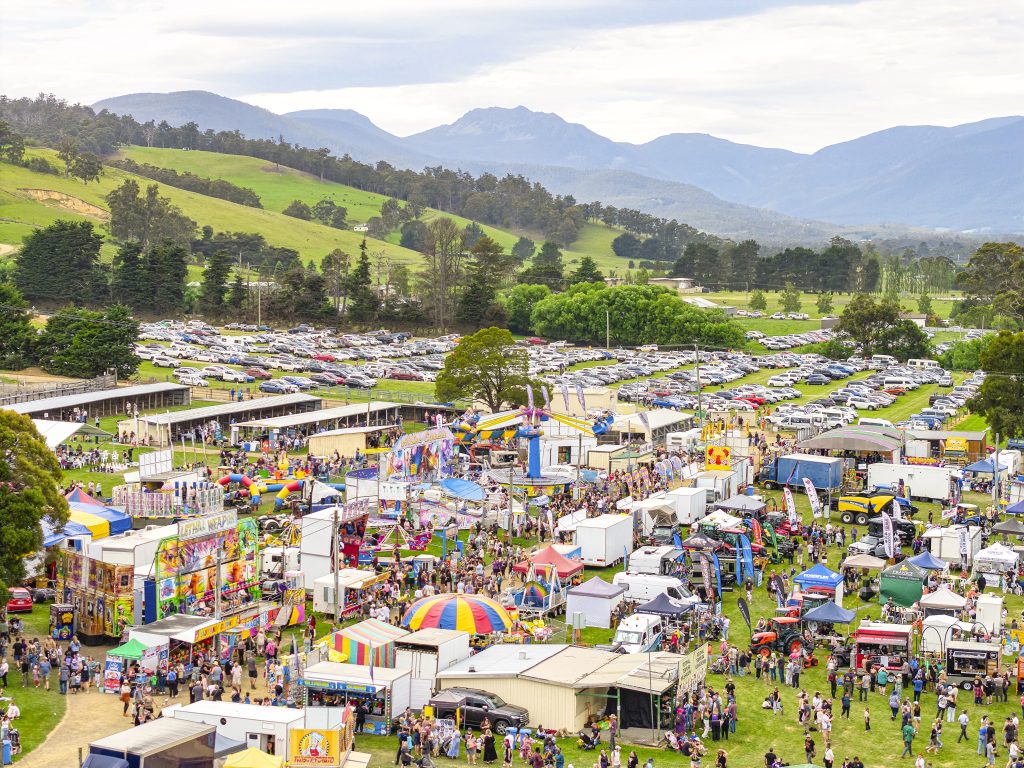 An outdoor fair with colorful stalls, rides, and crowds of people, set in a grassy field with parked cars and green hills in the background under a cloudy sky.