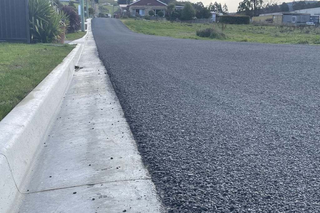 Freshly paved asphalt road with a concrete curb, leading uphill toward houses and greenery in the background. The sky is clear, and the area appears to be a quiet suburban or rural neighborhood.