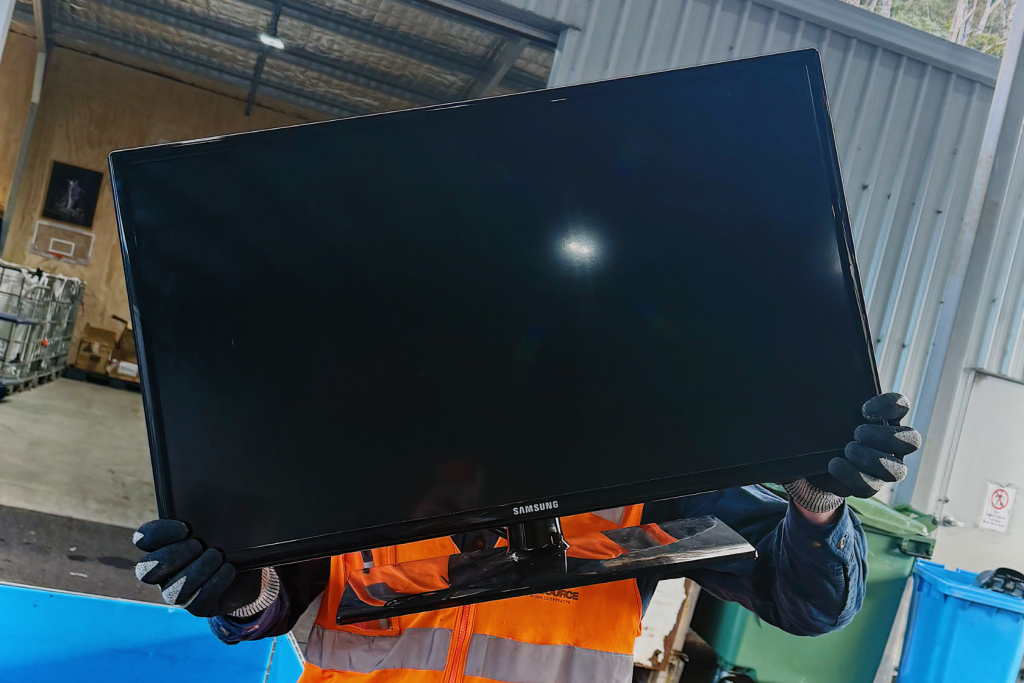 A person in orange safety gear holds a large Samsung flat-screen TV in front of them at a recycling or disposal facility.