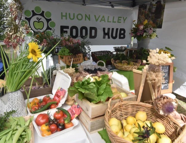 A market stall with fresh vegetables, fruits, flowers, and signs, under a banner reading HUON VALLEY FOOD HUB.