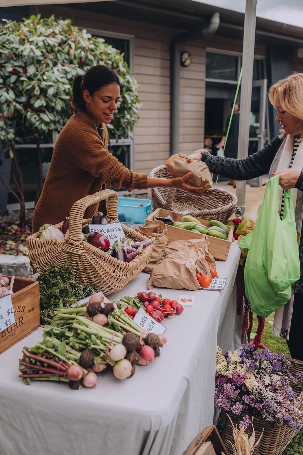 Two women exchange goods at an outdoor farmers market stall with baskets of fresh vegetables, radishes, eggplants, flowers, and produce displayed on a white tablecloth.