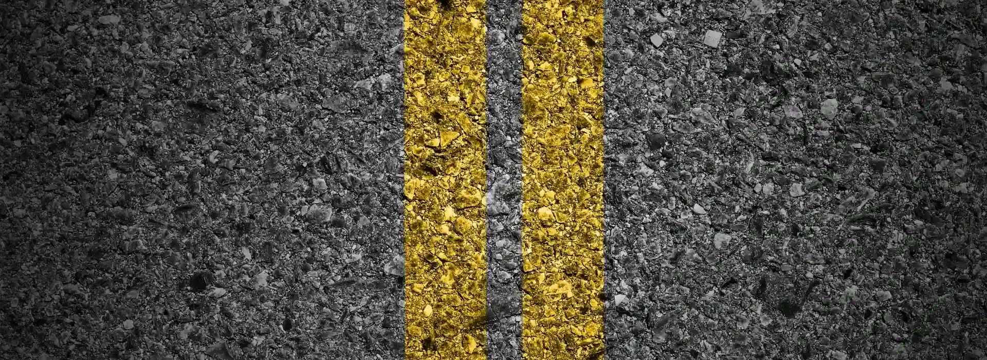 Close-up view of two yellow parallel lines painted on dark gray asphalt, representing road markings. The surface texture of the asphalt is rough and detailed.