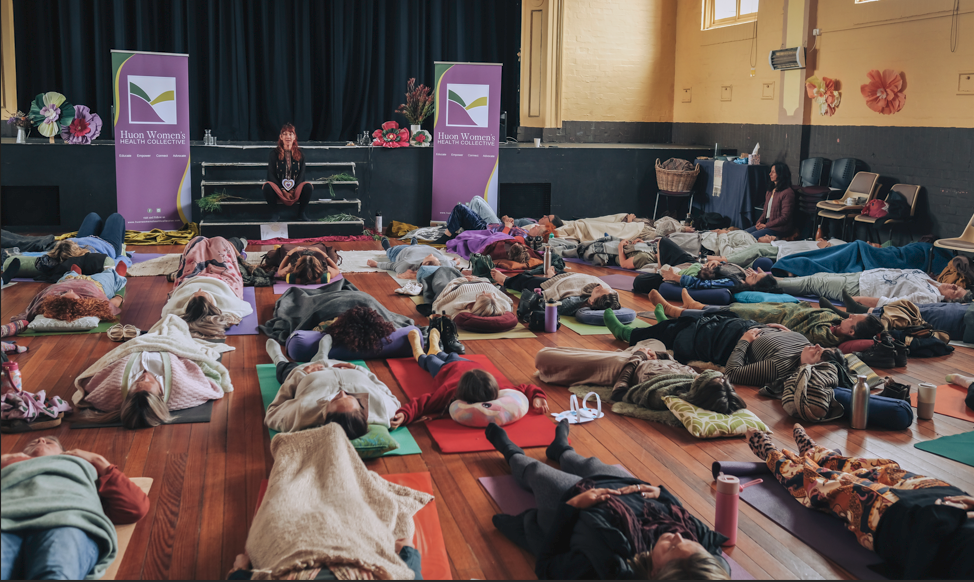 A group of people lies on yoga mats under blankets in a hall, with a woman seated on a stage in front of two banners. The atmosphere appears calm and meditative.