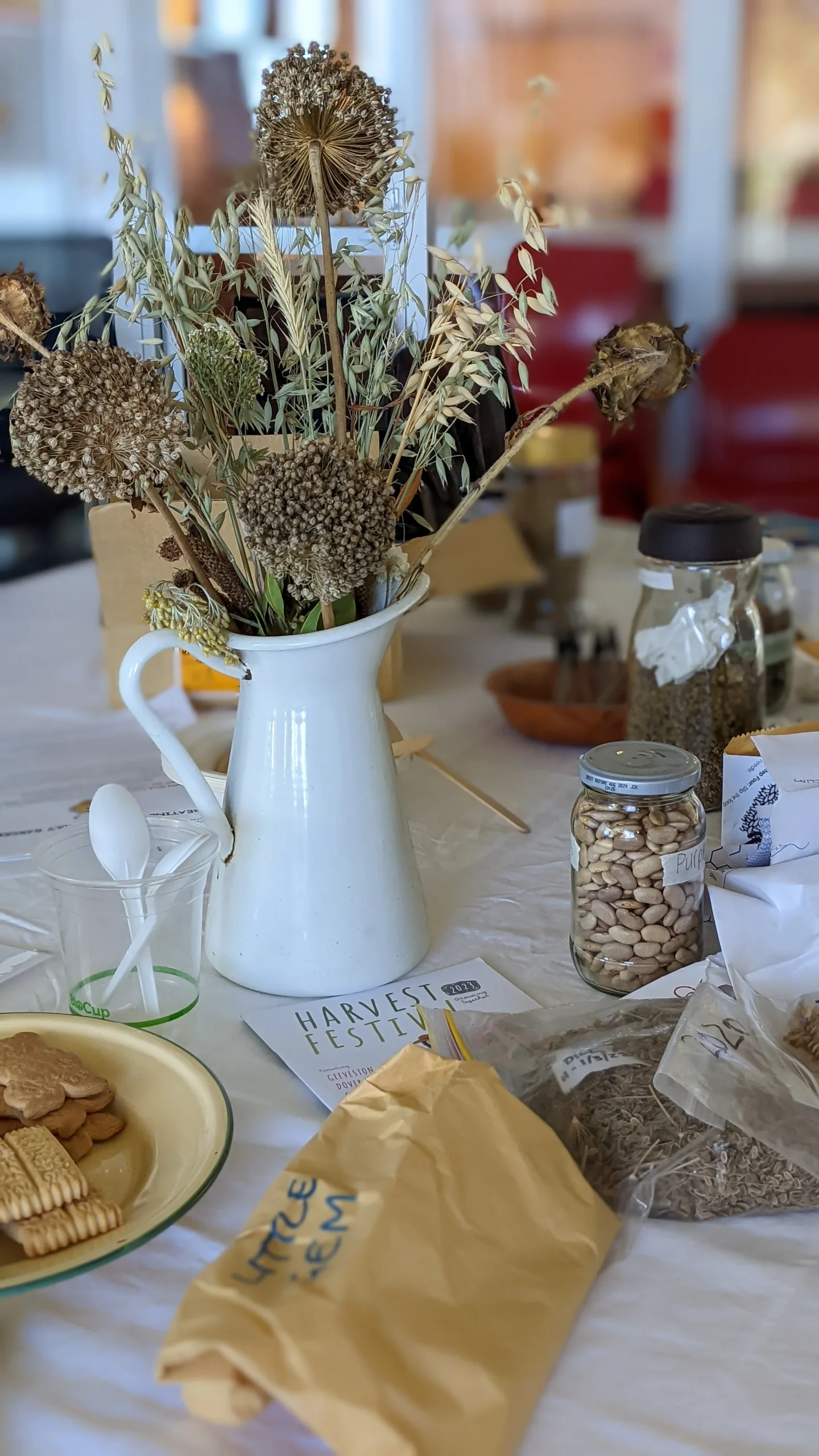 A white pitcher with dried flowers sits on a table surrounded by jars of beans and seeds, plates of cookies, paper bags, spoons, and seed packets, creating a rustic, harvest-themed display.