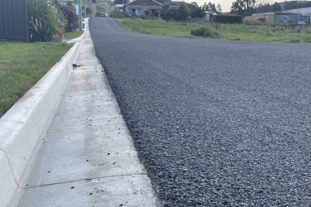 A newly paved asphalt road with a concrete curb, leading uphill toward houses in the distance.