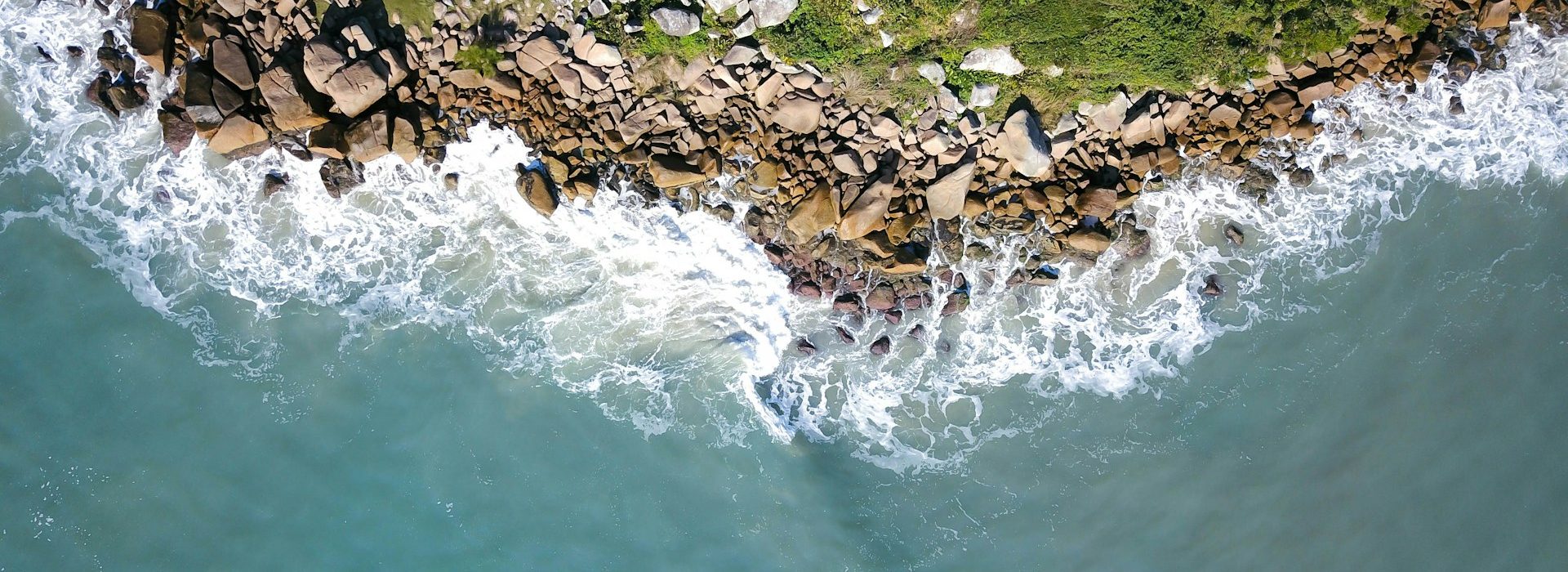 Aerial view of rocky coastline with waves crashing against the rocks and green vegetation on land. The ocean water appears blue-green and foamy near the shore.