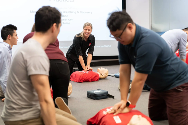 A group of adults practice CPR on training mannequins as an instructor guides them in a classroom setting.
