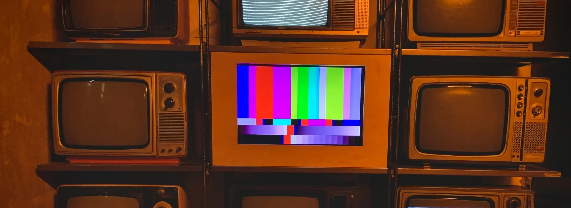 A stack of nine retro televisions, with one in the center displaying colorful test bars while the others show static or are turned off.