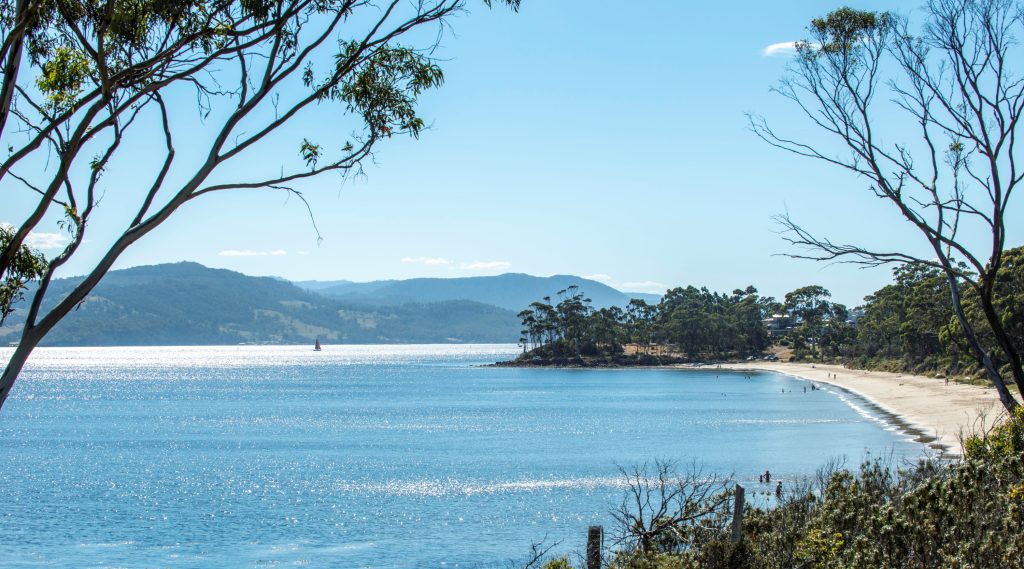 A calm beach with gentle waves, bordered by trees and hills under a clear blue sky. People walk along the sandy shore.